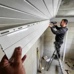 Un homme sur une échelle visse une lame de lambris PVC au plafond. Le chantier progresse.