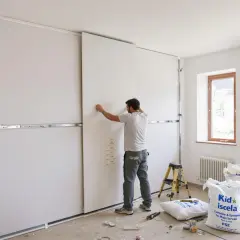 Un homme est en train de poser du placo sur une structure métallique dans une pièce en construction. Des sacs de matériaux sont au sol.