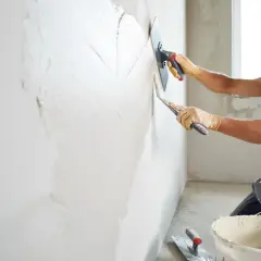Un ouvrier en tenue de travail, lunettes de protection et gants, est en train de faire de l'enduit sur un mur avec une taloche et une truelle.