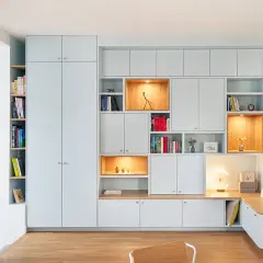 Un mur habillé de rangements modulables bleu pâle, avec des niches éclairées et une banquette intégrée près d'un escalier en bois.
