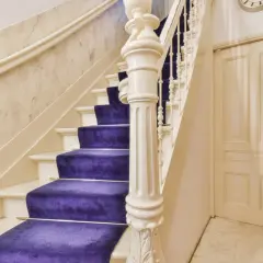 Escalier majestueux avec une moquette violette éclatante, un garde-corps blanc sculpté et un mur en marbre. Une porte crème et un mur de briques complètent la scène.