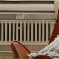 Un fauteuil en cuir marron avec un plaid douillet est placé près de vieux panneaux rayonnants blancs, créant une ambiance chaleureuse.