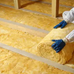 Pose d'isolant jaune, peut-être laine de verre vs laine de roche, dans un grenier. Mains gantées bleues déroulant un rouleau.