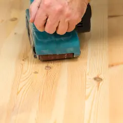 Main d'une personne utilisant une ponceuse pour poncer un parquet en bois clair.