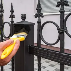 Maintenir le fer forgé propre avant peinture. Une main tient un pinceau jaune pour nettoyer la clôture.