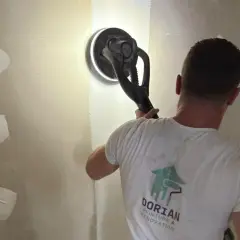 Un ouvrier utilise une ponceuse pour lisser un mur, préparant la surface pour poser bande placo.