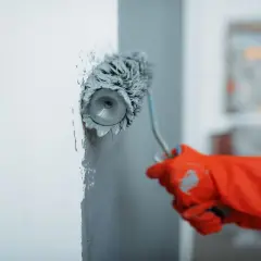Maintenant, vous savez comment peindre un mur : une main gantée de rouge utilise un rouleau pour appliquer de la peinture grise sur un mur blanc.