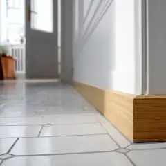 Pose de plinthe en bois clair, finition impeccable sur un sol carrelé blanc. La lumière du jour met en valeur le travail.