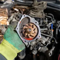 Main d'un mécanicien tenant une pompe à eau électrique démontée, prête pour la réparation. Le moteur est visible en arrière-plan.