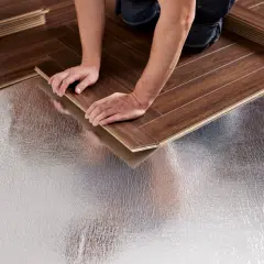 Pose bâton rompu : des mains ajustent une lame de parquet sur une sous-couche isolante argentée.