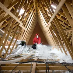 Un ouvrier en masque et sweat rouge réalise l'isolation des combles par soufflage, projetant de la ouate de cellulose dans la charpente.