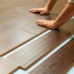Pose de lames de parquet imitation bois sur une sous-couche bleue. L'isolation plancher bois est en cours, les mains guident l'assemblage.