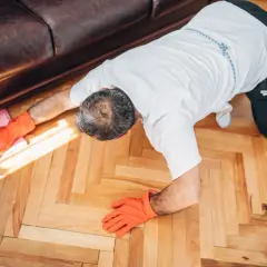 Un homme, vêtu d'une chemise blanche et de gants orange, est à genoux sur un parquet en bois. Il semble apprendre comment cirer un parquet, utilisant une lingette rose.