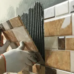 Mains gantées appliquant du mortier pour la pose carrelage d'une mosaïque murale aux tons marron et beige.