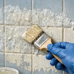 Main bleue appliquant un produit pour enlever peinture sur carrelage bleu décrépit. Le produit dissout la peinture par endroits.