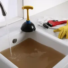 Évier bouché avec de l'eau sale. Un déboucheur, une clé à molette rouge et des gants jaunes sont prêts pour l'action.