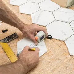 Mains mesurant une tuile hexagonale blanche sur un plancher OSB, avec des outils de pose.