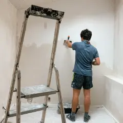 Un homme enlever du crépi intérieur sur un mur, à l'aide d'une spatule, près d'un escabeau.