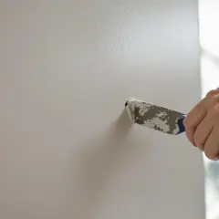 Main tenant utilise un couteau à mastic pour reboucher un trou dans un mur blanc.