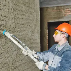 Un ouvrier en casque orange utilise un pistolet à crépir pour étanchéifier un mur en béton brut.