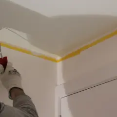 Main tenant en main un rouleau, il s'apprête à peindre un plafond. Le ruban de masquage jaune protège les bords.