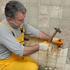 Un homme en salopette jaune utilise un marteau pour enlever du carrelage mural.