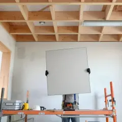 Un ouvrier sur un échafaudage s'apprête à poser une plaque pour faire un faux plafond. La structure en bois est visible.