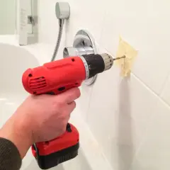Une main utilise une perceuse pour percer du carrelage dans une salle de bain, avec du ruban adhésif pour guider le foret.