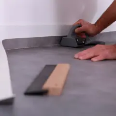 Pose lino : un artisan utilise un outil pour bien aplatir le revêtement de sol gris près du mur.