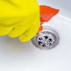 Gant jaune versant des granulés dans un siphon de douche pour déboucher la douche.