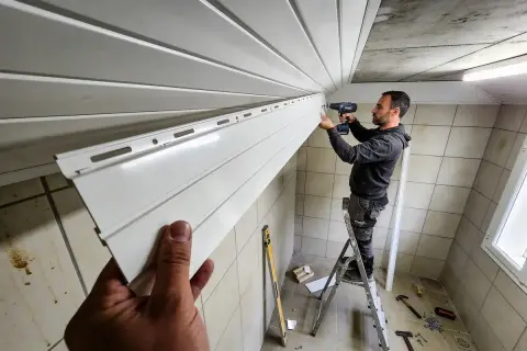 Un homme sur une échelle visse une lame de lambris PVC au plafond. Le chantier progresse.