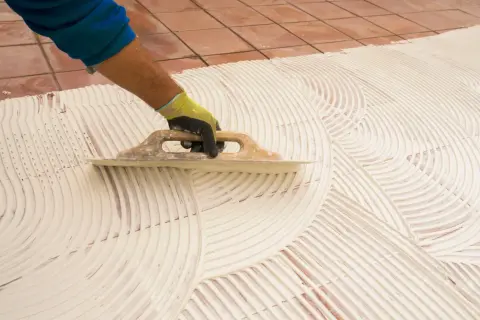 Main tenant, pose carrelage 60x60. Un artisan étale la colle avec une truelle pour préparer la pose de grands carreaux.