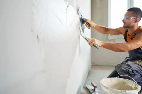 Un ouvrier en tenue de travail, lunettes de protection et gants, est en train de faire de l'enduit sur un mur avec une taloche et une truelle.