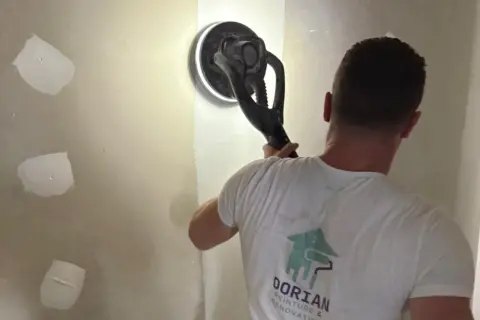 Un ouvrier utilise une ponceuse pour lisser un mur, préparant la surface pour poser bande placo.