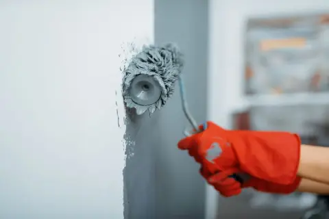 Maintenant, vous savez comment peindre un mur : une main gantée de rouge utilise un rouleau pour appliquer de la peinture grise sur un mur blanc.