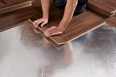Pose bâton rompu : des mains ajustent une lame de parquet sur une sous-couche isolante argentée.