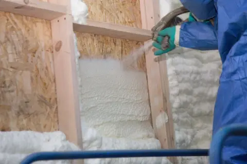 Application de polyuréthane projeté pour isoler une structure en bois. La mousse blanche s'étend, créant une barrière isolante.