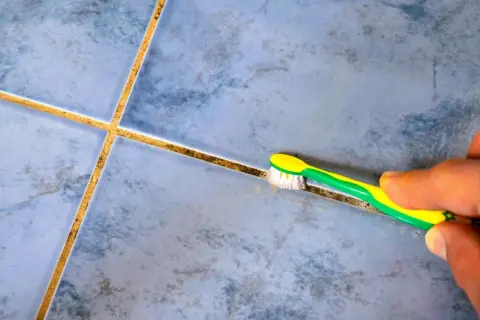 Main tenant, une brosse à dents verte et jaune est utilisée pour nettoyer joint salle de bain.