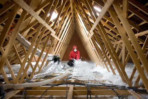 Un ouvrier en masque et sweat rouge réalise l'isolation des combles par soufflage, projetant de la ouate de cellulose dans la charpente.