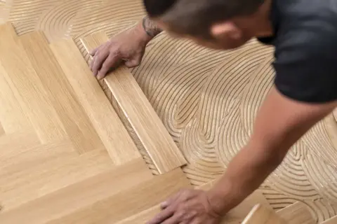 Pose d'un parquet flottant en chevrons. Un artisan applique la colle sur le sol avant de placer les lames de bois.