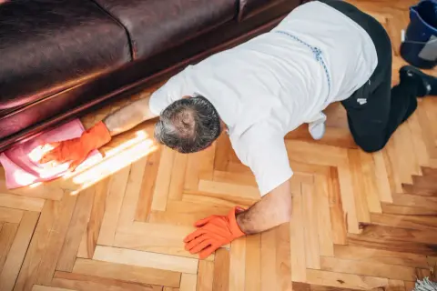 Un homme, vêtu d'une chemise blanche et de gants orange, est à genoux sur un parquet en bois. Il semble apprendre comment cirer un parquet, utilisant une lingette rose.