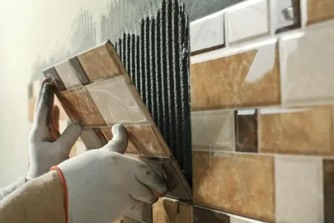 Mains gantées appliquant du mortier pour la pose carrelage d'une mosaïque murale aux tons marron et beige.