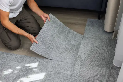 Pose d'une nouvelle moquette en dalles grises, recouvrant un ancien sol. Un homme pose une dalle, le geste est précis, comme s'il s'agissait de carreler sur du carrelage.