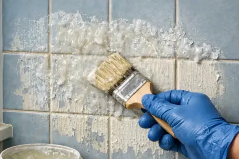 Main bleue appliquant un produit pour enlever peinture sur carrelage bleu décrépit. Le produit dissout la peinture par endroits.