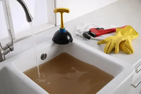 Évier bouché avec de l'eau sale. Un déboucheur, une clé à molette rouge et des gants jaunes sont prêts pour l'action.