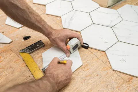 Mains mesurant une tuile hexagonale blanche sur un plancher OSB, avec des outils de pose.