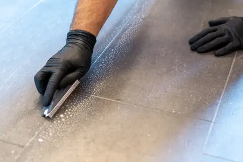Main tenant enlever joint carrelage avec une spatule. Des bulles de savon indiquent le nettoyage.