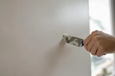Main tenant utilise un couteau à mastic pour reboucher un trou dans un mur blanc.