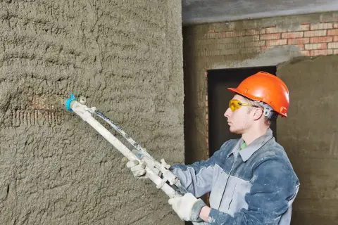 Un ouvrier en casque orange utilise un pistolet à crépir pour étanchéifier un mur en béton brut.