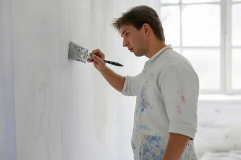 Un peintre, vêtu d'une chemise tachée, applique une couche de peinture sur un mur déjà peint, esquissant un portrait.